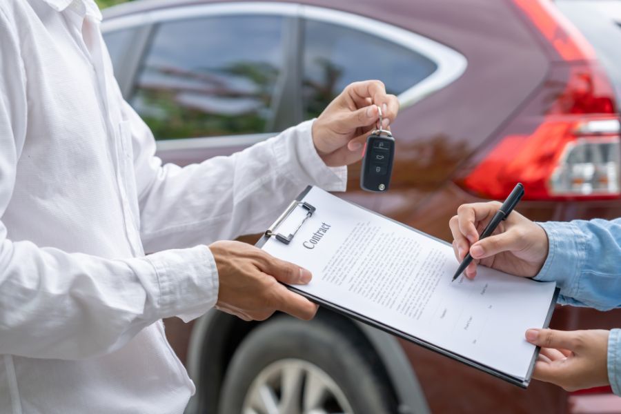 Un agent en chemise blanche tenant des clés de voiture pendant qu'un client signe un contrat de location de voiture devant un véhicule rouge.